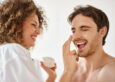 Woman applying cream to her man in bathroom and laughing together, cheerful happy couple - Skincare for men and women