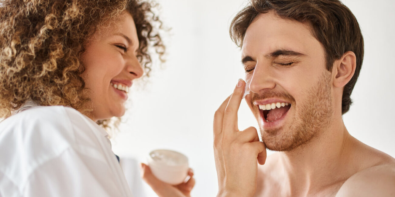 Woman applying cream to her man in bathroom and laughing together, cheerful happy couple - Skincare for men and women