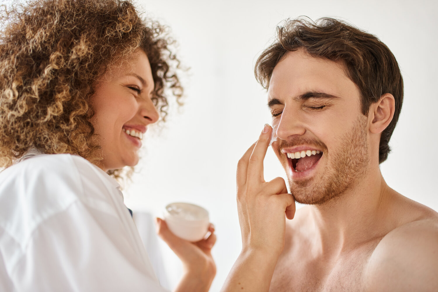 Woman applying cream to her man in bathroom and laughing together, cheerful happy couple - Skincare for men and women